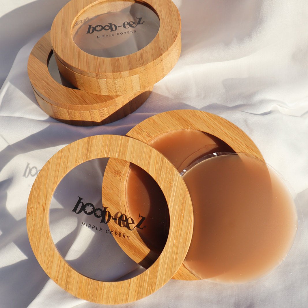 Product shot of a Boob-eez Bamboo Compact with Dark tint Headlight Hiders inside with Bamboo Compacts and nipple covers in the background, close-up shot showing the beautiful wood grain of the bamboo and the magnetic closure
