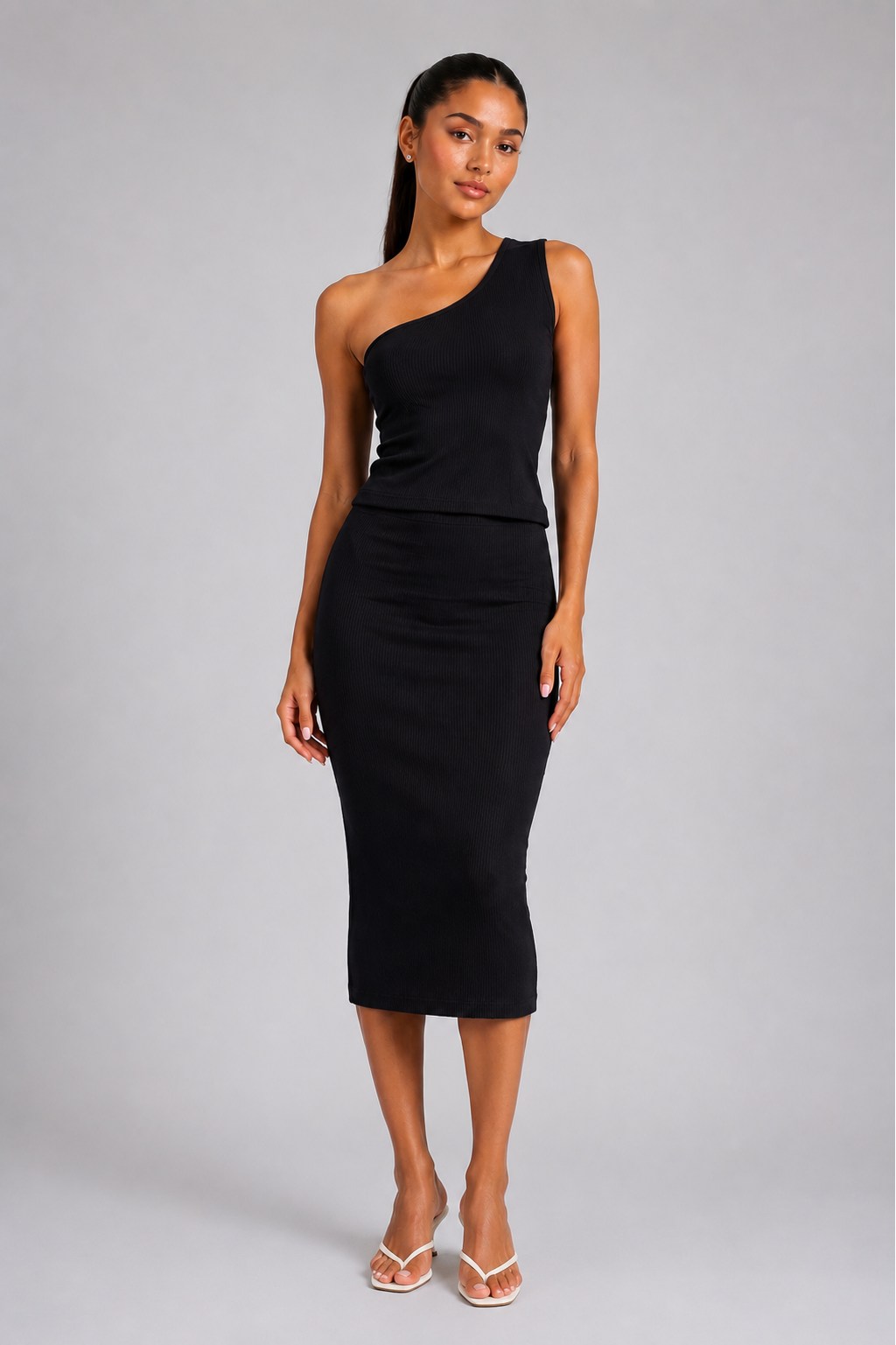 A woman modeling a black one-shoulder midi dress with a peplum waist, paired with white strappy sandals, against a solid gray studio background.