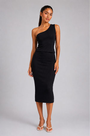 A woman modeling a black one-shoulder midi dress with a peplum waist, paired with white strappy sandals, against a solid gray studio background.