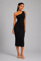 A woman modeling a black one-shoulder midi dress with a peplum waist, paired with white heeled sandals, against a solid gray studio background.