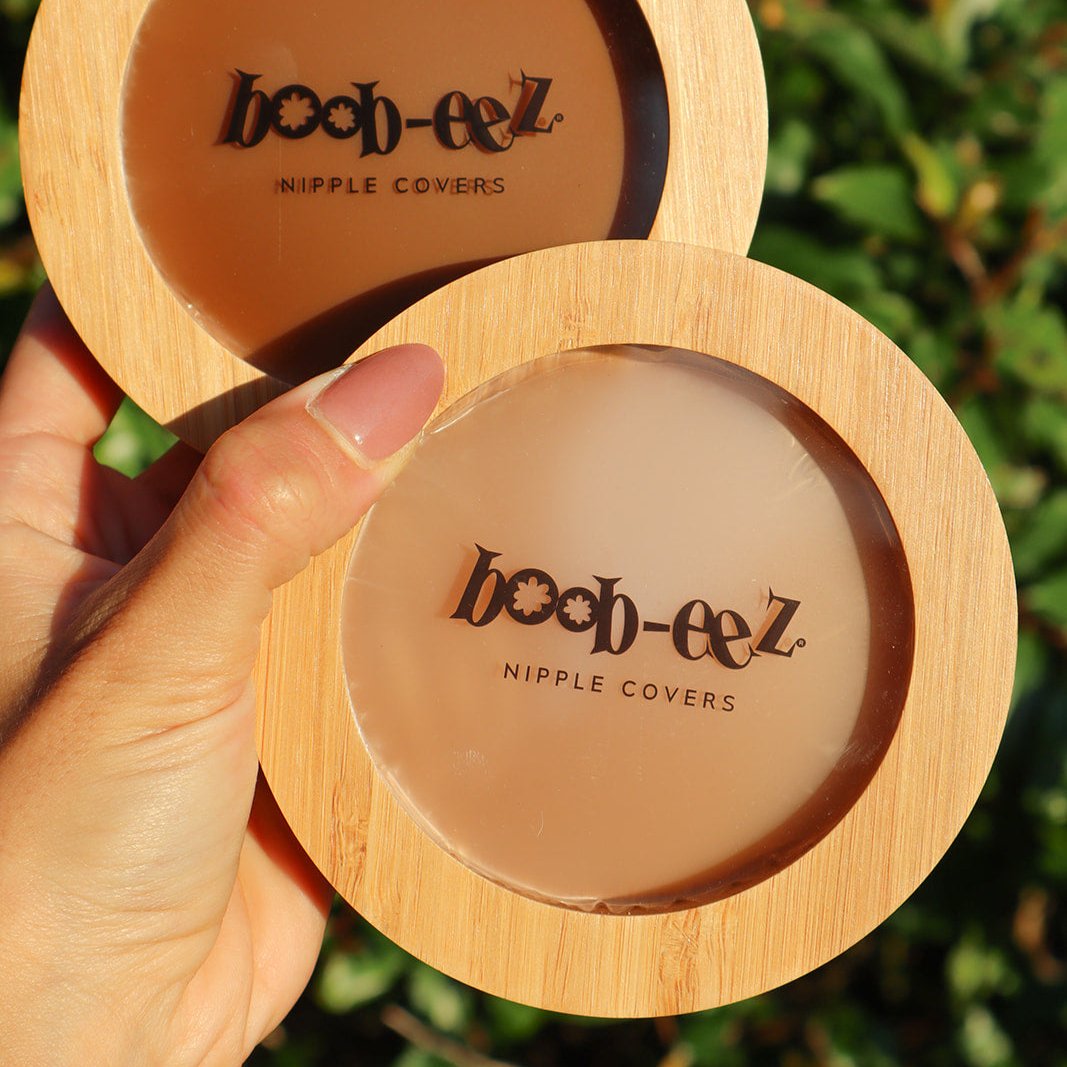 Product shot of a hand holding a Boob-eez Bamboo Compact with Dark tint Headlight Hiders inside and a Bamboo Compact with Medium tint nipple covers inside with natural green in the background, close-up shot showing the beautiful wood grain of the bamboo and the transparent window to show the sticky nipple covers inside
