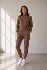 A woman modeling a matching brown lounge set consisting of a half-zip sweatshirt and jogger pants, paired with white sneakers, in a minimalist room with window shadows.