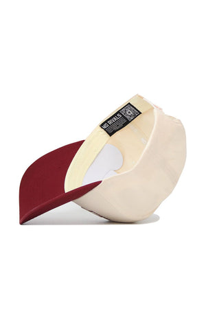 The Montana University Triangle Game Cap Hat by No Rivals is shown upside down, revealing its inside labels and stitching. The beige snapback features a maroon brim, with "NO RIVALS" noted on the interior label against a plain white background.