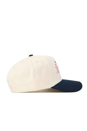 The No Rivals DBU Triangle Game Cap Hat is a cream-colored, structured fit baseball cap with a navy blue brim and partial red embroidery on the front, shown from the side. Officially licensed.
