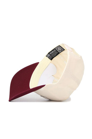 The New Mexico State Triangle Game Cap Hat by No Rivals is shown upside down, displaying its cream color, maroon brim, white sweatband, and a black tag with text and a QR code inside.