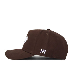The Liberty Americano Hat is a brown, structured cotton cap with a curved brim. It features white embroidered NR letters on the side, a partially visible front logo, and is shown from a side view against a white background.