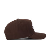 The Liberty Americano Hat is a brown structured-fit cap crafted from high-quality cotton, shown side-on with visible stitching, a slightly curved brim, and a partial white embroidery on the front. The background is plain white.