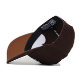 The Liberty Americano Hat, a brown structured fit snapback made from high-quality cotton, is shown on its side, highlighting the curved brim and interior labels that read NO RIVALS.