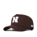The Nebraska Americano Hat is a structured brown cotton cap with a large white "N" on the front, "NR" on the side, a curved brim, and an adjustable back strap.