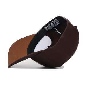 The University of Montana Americano Hat by No Rivals is a structured brown snapback made from high-quality cotton, featuring an inside label with "NO RIVALS" and a QR code on the black adjustable strap.