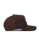 The Georgia Americano Hat, a brown snapback with a structured fit, is shown from the side on a white surface. Made of high quality cotton, it has a curved brim and features a circular patch on the front, not fully visible here.