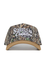 The Scheels Camo Explorer Hat is a camouflage baseball cap with a tan brim, structured fit, snapback closure, white “Scheels Outfitters Since 1902” embroidery, and two tree graphics on the front.
