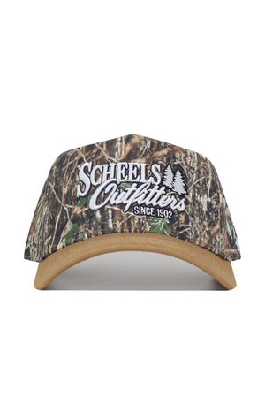 The Scheels Camo Explorer Hat is a camouflage baseball cap with a tan brim, structured fit, snapback closure, white “Scheels Outfitters Since 1902” embroidery, and two tree graphics on the front.