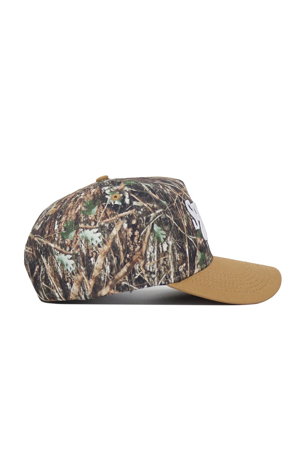 The Scheels Camo Explorer Hat is shown from the side on a white background, featuring a brown camo-patterned crown with grass and leaves, solid tan brim, structured fit, and high-quality cotton construction.