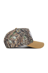 The Scheels Camo Explorer Hat is shown from the side on a white background, featuring a brown camo-patterned crown with grass and leaves, solid tan brim, structured fit, and high-quality cotton construction.