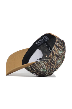 The Scheels Camo Explorer Hat is shown on its side, highlighting the inside label and black sweatband. It features a brown brim and a crown with a green, brown, and tan camo pattern.