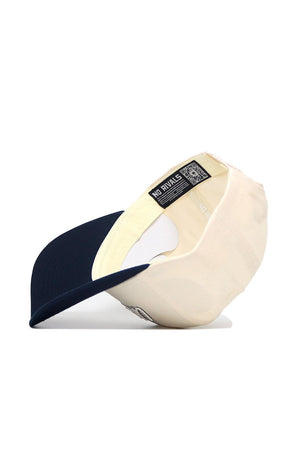 The UConn Triangle Game Cap Hat, cream-colored with a navy brim, is shown upside down to display its quality cotton interior, a NO RIVALS label, and an inner band QR code for authenticity.