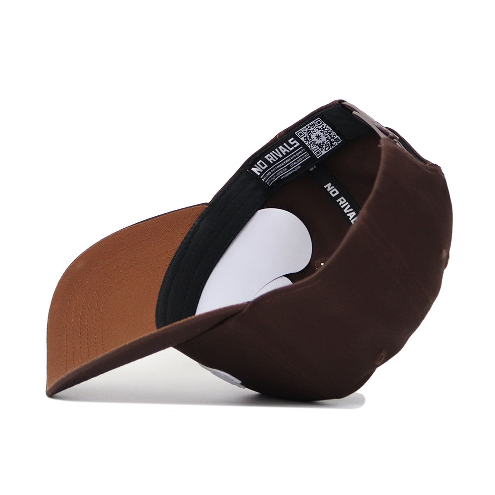 The JMU Americano Hat, a brown structured-fit cotton snapback, is shown from the back and underneath. Its inside label reads NO RIVALS next to a QR code. The hat is placed on its side against a white background.