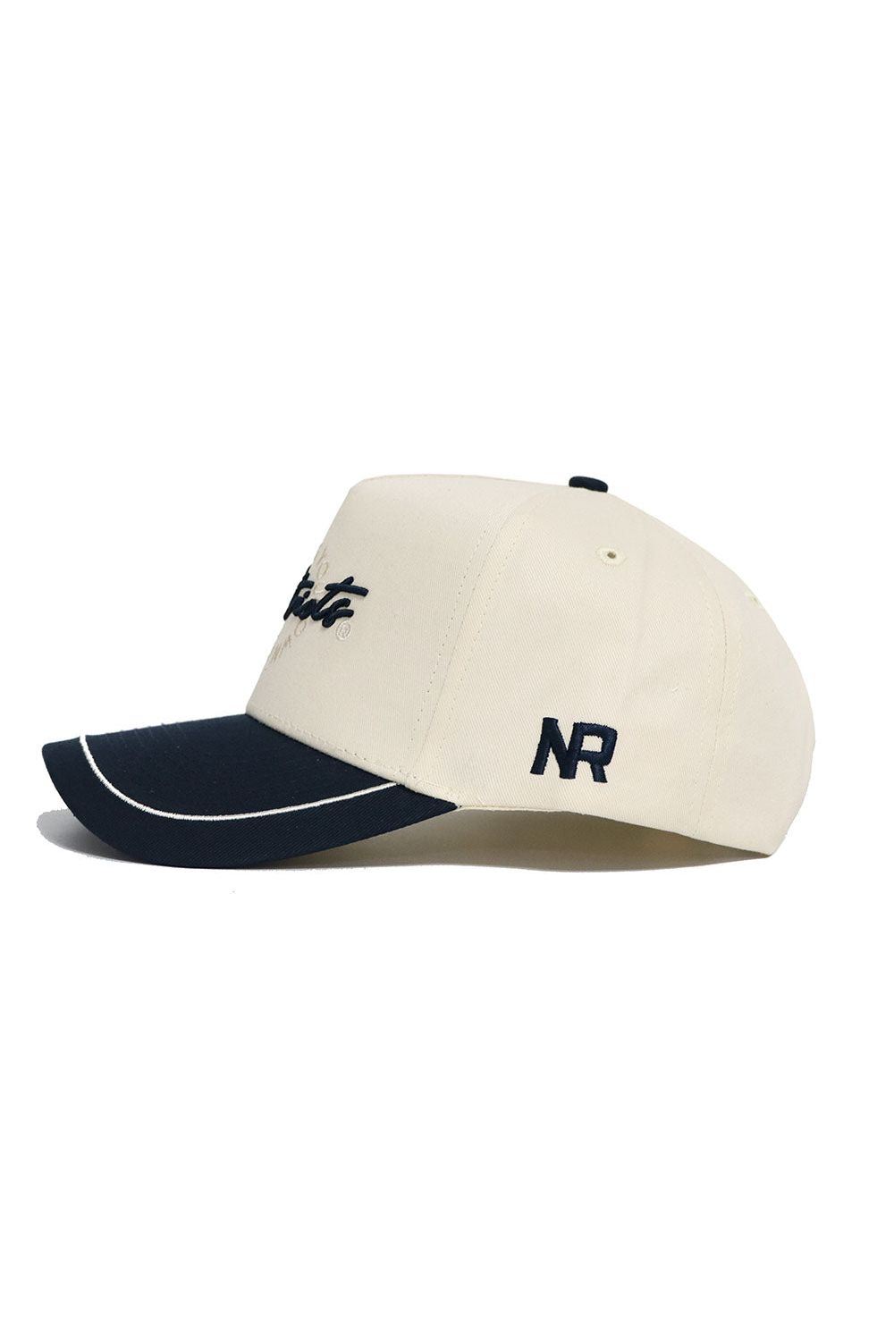 The DBU Patriots ALOYO Hat by No Rivals is a cream snapback with a black curved brim, white trim, and high-quality cotton. It features "NR" stitched in black on the side and partially visible script embroidery on the front for a structured fit.