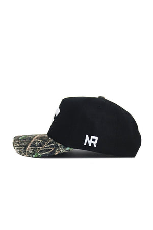 The K-State Camo Hat - Trailblazer Edition by No Rivals features a black snapback design, a camo-patterned brim, and white NR embroidery on the side. Made from high-quality cotton, it's shown from the side against a white background.