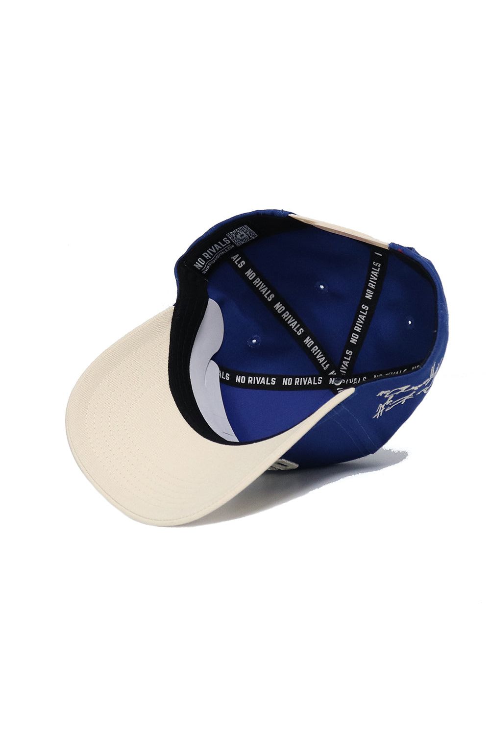The OWLS Established Hat by No Rivals is shown upside down, displaying its blue and beige inner lining and “NO RIVALS” on the inside band. White stitching and a side signature add standout style—ideal for your gameday look.