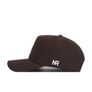 The Wyoming Americano Hat is a brown cotton baseball cap shown from the side, with white embroidered "NR" on the side panel. It features a curved brim, ventilation eyelets, and a classic snapback closure.