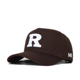 The Rutgers Americano Hat (B2B) is a brown cotton snapback with a large white "R" on the front, "NR" stitched in white on the side, a curved brim, and structured fit, shown against a white background.