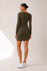 A woman wearing a fitted long-sleeve mini dress in olive green, shown from the back against a light neutral studio background. She is also wearing white sneakers.