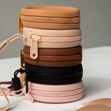 Product shot of a stack of reusable Silicone Travel Cases for Headlight Hiders in Medium Nude, Light Nude, Dark Nude, Dusty Pink, and Black, showing the zipper detail and removable strap