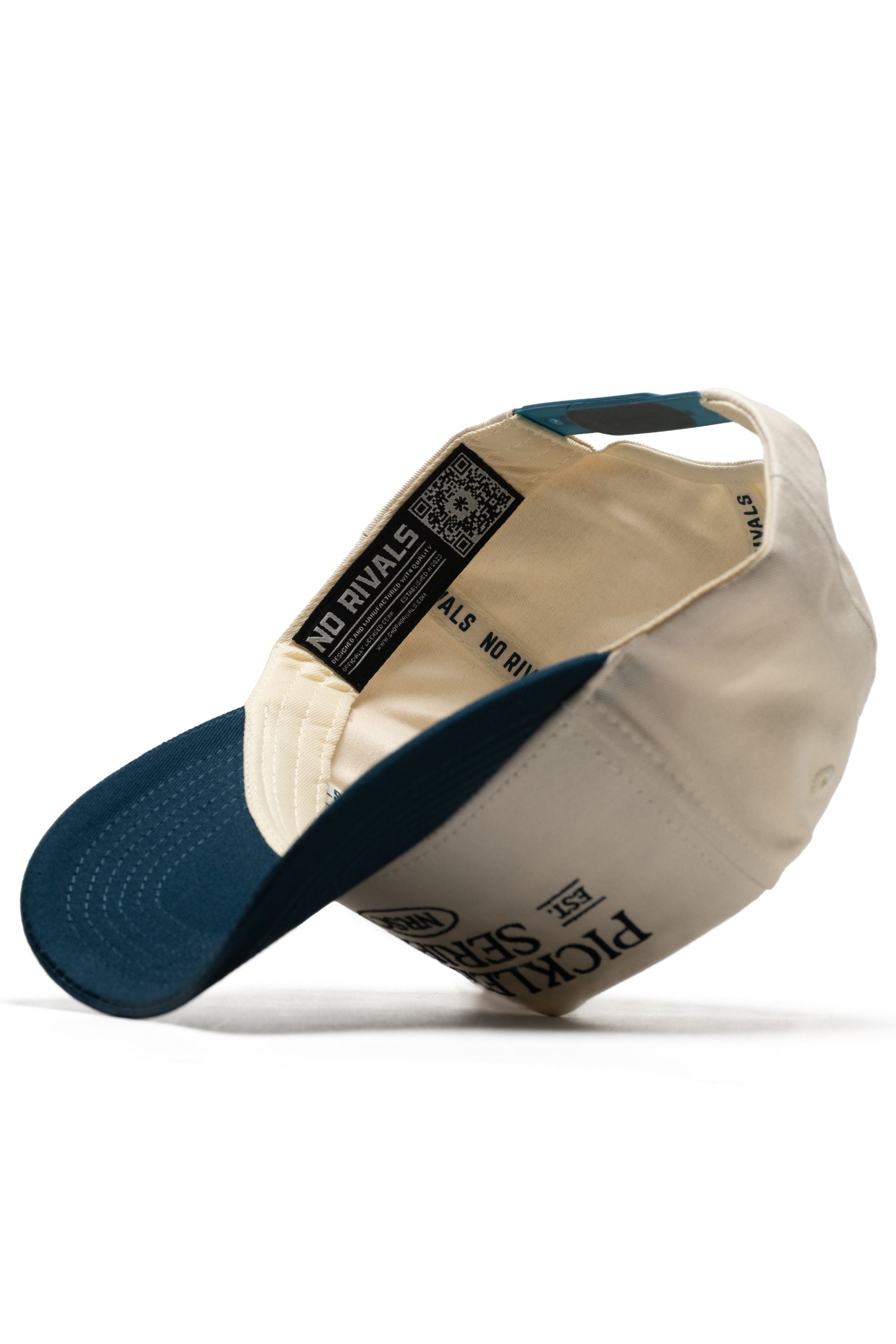 The Old School Pickleball Hat - Classic Blue by No Rivals is shown inside out, displaying a branded label, QR code on the inner band, visible stitching, structured fit, and premium cotton construction in cream and navy blue.