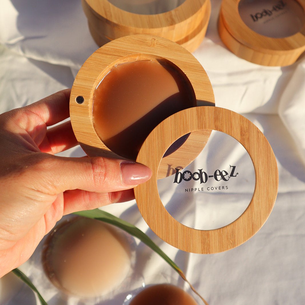 Product shot of a hand holding a Boob-eez Bamboo Compact with Dark tint Headlight Hiders inside with Bamboo Compacts and nipple covers in the background, close-up shot showing the beautiful wood grain of the bamboo and the magnetic closure