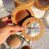 Product shot of a hand holding a Boob-eez Bamboo Compact with Dark tint Headlight Hiders inside with Bamboo Compacts and nipple covers in the background, close-up shot showing the beautiful wood grain of the bamboo and the magnetic closure