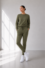 A woman modeling a matching olive green lounge set consisting of a long-sleeve crewneck sweatshirt and drawstring jogger pants, paired with white sneakers in a bright, modern room with window light.