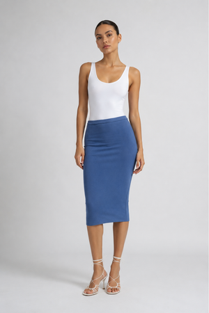 A model stands against a light gray studio background wearing a white sleeveless top and a fitted blue midi skirt, paired with clear high-heeled sandals.