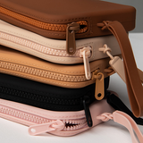Product shot of a stack of reusable Silicone Travel Cases for Silicone Stick-on Bra in Dark Nude, Light Nude, Medium Nude, Black, and Dusty Pink, showing the zipper detail and removable strap