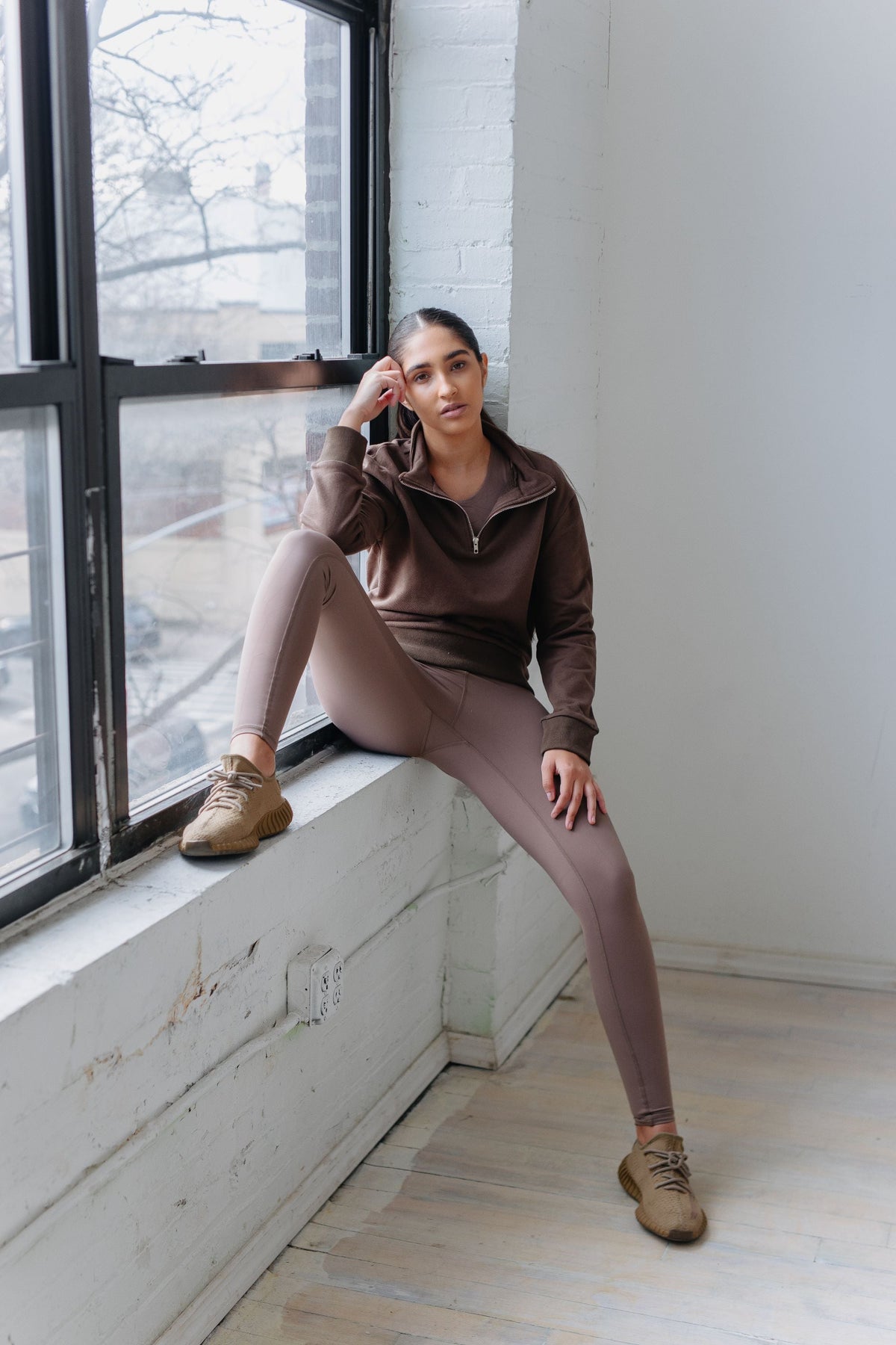 Woman in brown tracksuit sitting by a window in a minimalistic room.