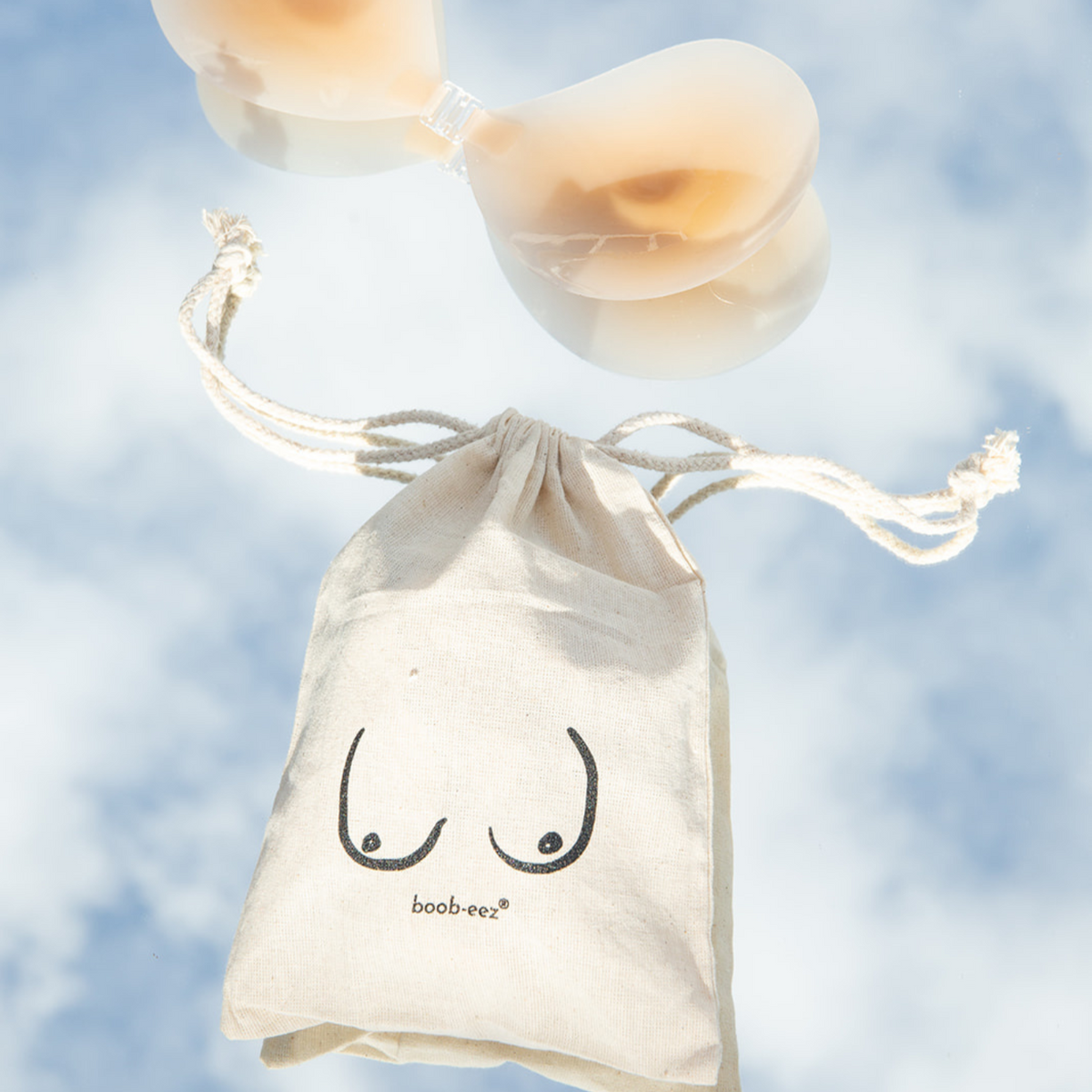 Product shot of Silicone Stick-on Bra with Boob-eez branded drawstring unbleached cotton muslin pouch laying flat against mirrored sky background showing how thin and weightless the Silicone Stick-on Bra is 